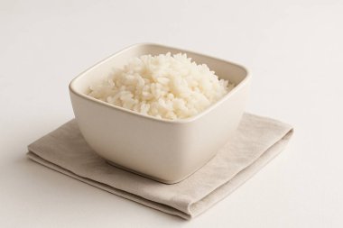 Bir fotoğraf, bej bir peçeteye yerleştirilmiş pişmiş beyaz pirinçle dolu kare bir seramik kaseyi sergiliyor. Yumuşak ışıklandırma ve nötr arkaplan ile minimalist kompozisyon.