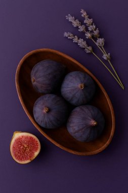 Yumuşak mavi-mor bir zemin üzerinde, ahşap oval bir kâse içinde olgun incir üst aşağı görünüm, bir incir yarı aşağıda ve hassas bir dal üzerinde. Minimalist kompozisyon.