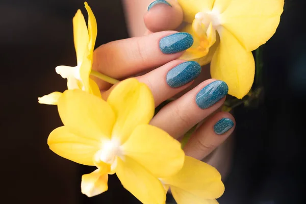 Female hand with beautiful manicure - blue glitter nails with yellow phalaenopsis orchid flower. Nail care concept