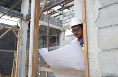 Kendine güvenen bir portre mühendisi inşaat alanındaki planlı turuncu yelek ve turuncu kask giyer. Profesyonel ve endüstriyel, mimarlık işi, fabrikada çalışan işçiler..