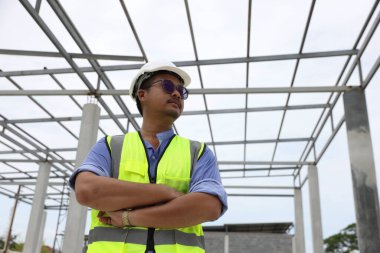 Kendine güvenen bir portre mühendisi inşaat alanındaki planlı turuncu yelek ve turuncu kask giyer. Profesyonel ve endüstriyel, mimarlık işi, fabrikada çalışan işçiler..