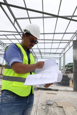 Kendine güvenen bir portre mühendisi inşaat alanındaki planlı turuncu yelek ve turuncu kask giyer. Profesyonel ve endüstriyel, mimarlık işi, fabrikada çalışan işçiler..
