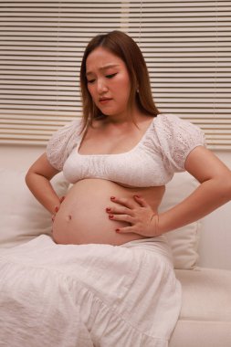 Hamilelik karnına yakın çekim. Hamile kadınlar beyaz bir elbise giyip evde rahatlıyor. Cilt bakımı, sağlık, yaşam tarzı, hamile kadınlar bebek bekliyor.