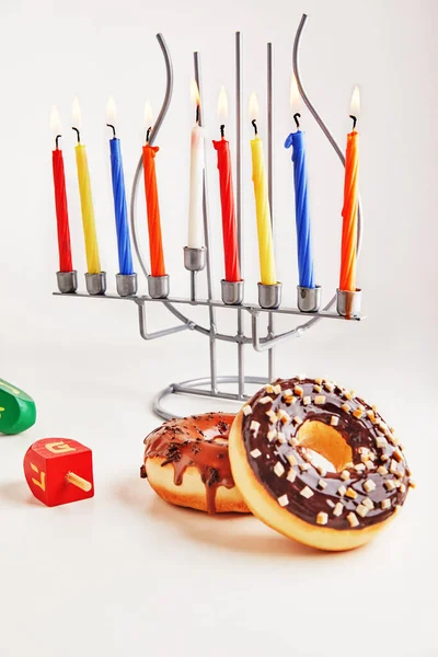 Jewish holiday Hanukkah background. Traditional dish is sweet donuts. Hanukkah table setting candlestick with candles and spinning tops on white background. Letters of Hebrew alphabet on tops. Copy space.