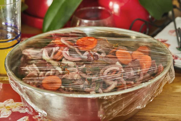 Diced beef with onions and carrots in metal bowl covered with cling film, ready to cook.