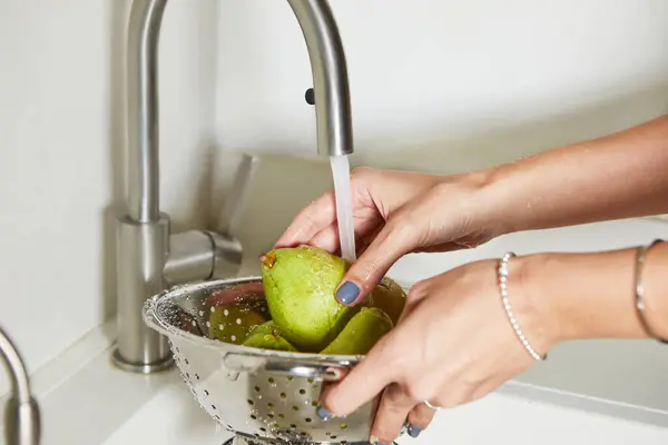 A woman is washing a handful of pears under running water in a kitchen sink. The pears are green and the water is clear. The woman is wearing a white shirt and the sink is made of stainless steel.
