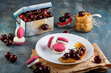 Masada rengarenk badem kurabiyeleri ve taze kirazlarla süslenmiş güzel beyaz bir tabak. Pişmiş yiyecekler ve meyvelerin enfes bir kombinasyonunu sergiliyor.