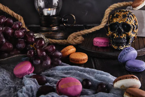 stock image A dramatic still life with red wine, dark grapes, colorful macarons, and an ornate skull decoration, set against a dark background.