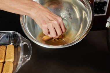 Bir kadın cam bir kasede tiramisu tatlısı hazırlıyor. Yanında da ev mutfağında karıştırma kabı var. Adım adım tarif.