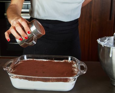 Ev yapımı tiramisu tatlısının üzerine kakao tozu tozu döken bir adam. Yanında karıştırma kabıyla birlikte..