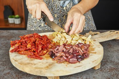 Yakın plan çekimde zeytin, kurutulmuş domates ve diğer malzemeler mutfakta ahşap kesim tahtalarında kesiliyor..
