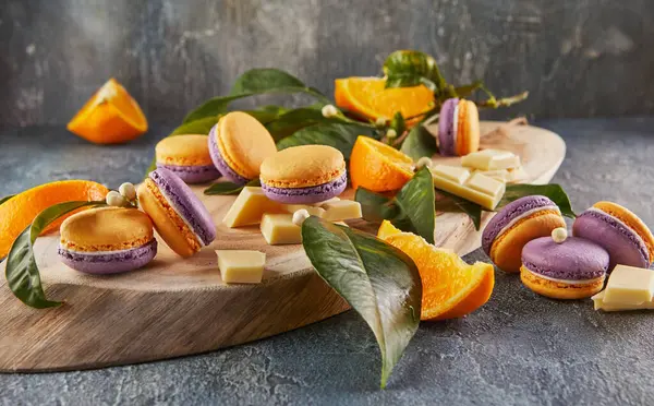 stock image Vibrant yellow and purple macarons displayed with orange slices, green leaves, and white chocolate chunks on a rustic wooden board.