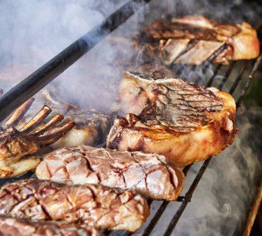 Sulu biftek ve pirzolaların dumanlı bir barbeküde ızgaraya dönüşmesi, açık hava ortamında char izleri ve cızırdayan etleri gösteriyor..