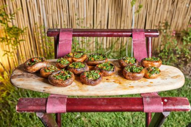 İnce doğranmış otlarla kaplı ızgara mantarlar, açık hava ortamında ahşap bir tahta üzerine dizilmiş..