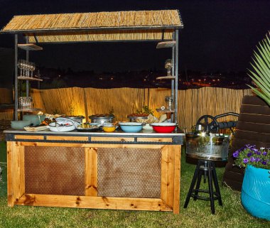 Bambu çatılı bir yapının altında çeşitli yiyecek ve içecek seçenekleri olan kırsal bir açık büfe tezgahı..