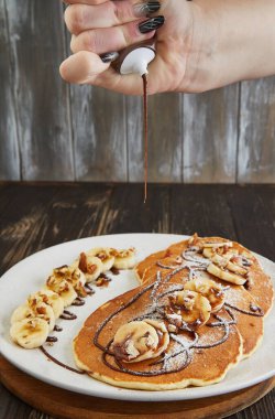 Close-up of a hand applying chocolate drizzle over pancakes topped with bananas and nuts, enhancing the dishs appeal and flavor. clipart