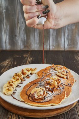 Close-up of a hand applying chocolate drizzle over pancakes topped with bananas and nuts, enhancing the dish s appeal and flavor. clipart