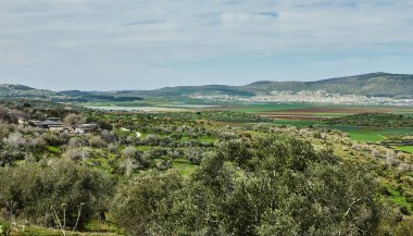 Parçalı bulutlu bir gökyüzünün altında yuvarlanan yeşil tepeler, zeytin ağaçları ve uzak tarım arazileri ile nefes kesici bir doğal manzara sükuneti çağrıştırıyor..