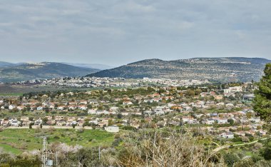 Vadideki bir kasabanın panoramik görüntüsü, yuvarlanan tepeler ve yemyeşil bulutlu bir gökyüzünün altında..