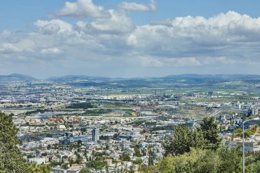 Endüstriyel bölgeleri, binaları ve gökyüzünün altında büyük bulutlarla benekli yeşillikleri olan geniş bir şehir manzarası..