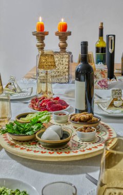 Akşam yemeği masası mumlarla aydınlatılmış geleneksel Hamursuz Bayramı yemekleri, şarap şişeleri ve bayramın törensel sembolizmi sergileniyor..
