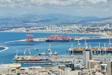Hayfa, İsrail - 13 Nisan 2025: Haifa limanı ve konteynır vinçleri, kargo gemileri ve bulutlu bir dağ zemininin altındaki konut binaları ile şehir silueti.