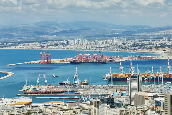 Hayfa, İsrail - 13 Nisan 2025: Haifa limanı ve konteynır vinçleri, kargo gemileri ve bulutlu bir dağ zemininin altındaki konut binaları ile şehir silueti.