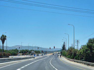 Trafik ışıkları ve açık gökyüzü ile geniş bir otoyol, yeşil ve uzak tepeler ile çevrili, parlak güneş ışığı altında.