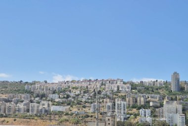 Yeşil yamaçlar boyunca yığılmış panoramik yerleşim binaları, mavi gökyüzü huzurlu bir şehir manzarası yaratıyor..