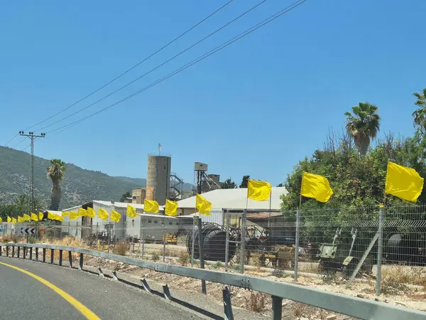 Sarı bayraklar, endüstriyel bir binanın yanındaki yol kenarındaki çitlerde palmiye ağaçları ve açık mavi gökyüzünün altındaki ormanlık tepeler..