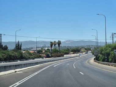 Modern bir asfalt yol yeşil ve çiçeklerin arasından kıvrılır, uzak tepeler tarafından çerçevelenir parlak ve bulutsuz bir yaz gökyüzü altında.
