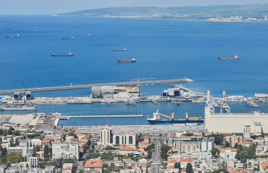 Park halindeki araçlar, liman terminalleri, kargo vinçleri ve ufukta uzanan derin mavi deniz ile Hayfa limanı ve kentsel ufuk çizgisi.
