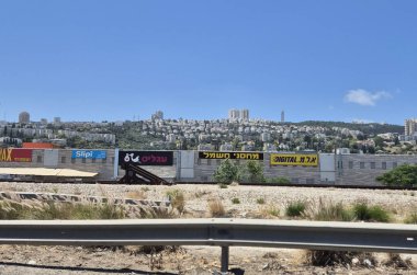 Hayfa, İsrail 24 Mayıs 2025: Ticari işaretler ve Haifa 'nın yol kenarından görülen uzak şehir manzarası, yukarısı konut binalarıyla dolu bir tepe.
