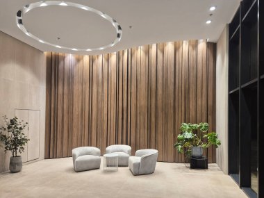 Modern bir lobide zarif kıvrımlı sandalyeler, ahşap panel duvarlar ve tavan ışıklarıyla aydınlatılmış dekoratif bitkiler bulunur..