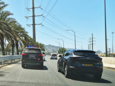 Hayfa, İsrail 24 Mayıs 2025: Bir polis aracı ve sivil araçlar liman yakınlarındaki palmiye çizgili bir yolda ilerlerken, şehir binaları uzak mesafeden görülebiliyor.