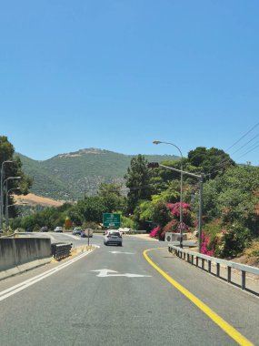 Hayfa, İsrail 24 Mayıs 2025: Kuzey İsrail 'de ağaçlarla kaplı bir yol ormanlar ve çiçeklerin arasından geçiyor, ileride trafik ışıklarına doğru giden arabalar var.