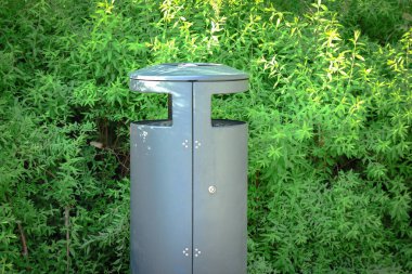 Metal trash can standing in green hedge foliage background in sunlight