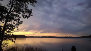 Sunset sky over lake landscape, nature background with heavy clouds and tree silhouette in front