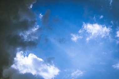 White soft clouds being covered by big dark cloud from left side while rays of light are coming from behind of it on blue sky