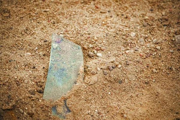 Transparent sharp glass shard from sticking out of the sand on the brown rural road with small pebbles