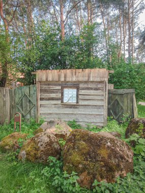 Küçük ahşap bir ev. Sahte penceresi var. Ağaçların arkasında, yosunlarla kaplı büyük bir kaya parçası.