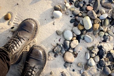 Sahilde kayalıklar ve çakıl taşlarıyla gün batımında tepeden aşağıya bakan kayalık botlar içinde ayakta duran bir kadının bacakları.