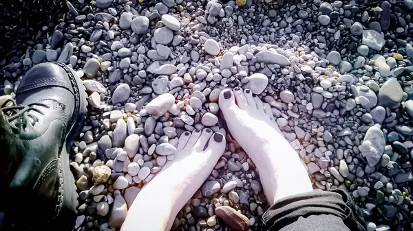 stock image Bare feet of a female rocker with black pedicure toes in pebbles with one rocker black boot standing near top down view