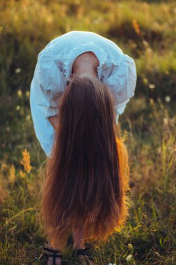 Güzel, uzun saçlı bir kadın saçını güneşli bir yaz tarlasında bağlamak için eğildi. Lüks, uzun, kırmızı, sağlıklı saçlar, doğal bakım ve bakım kavramı. Doğal gün batımı ışığı, seçici odak