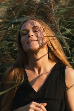 Siyah elbiseli, uzun kızıl saçlı, yeşil sazlıklara gülümseyen güzel bir kızın portresi. Doğada poz veren çekici kadın, seçici odaklanma.