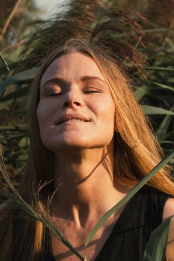 Siyah elbiseli, uzun kızıl saçlı, yeşil sazlıklara gülümseyen güzel bir kızın portresi. Doğada poz veren çekici kadın, seçici odaklanma.