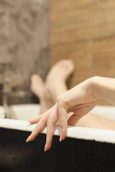 Beautiful female hand palm with natural manicure in foreground, legs in bath, lying relaxed, rest. SPA procedures for skin care, happy woman. Beautiful background, selective focus, close-up.