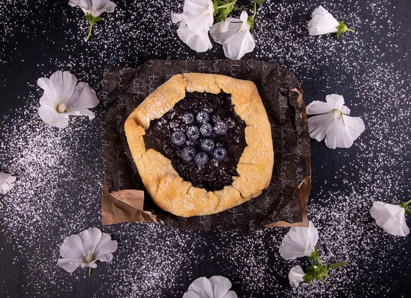 砂糖パウダーと白い花が並ぶ黒い背景にブルーベリーと夏の甘いパイケーキガレット トップビュー — ストック写真