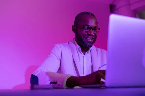 a black man in a white suit and glasses works at a laptop. a man in glasses works in the office in the night