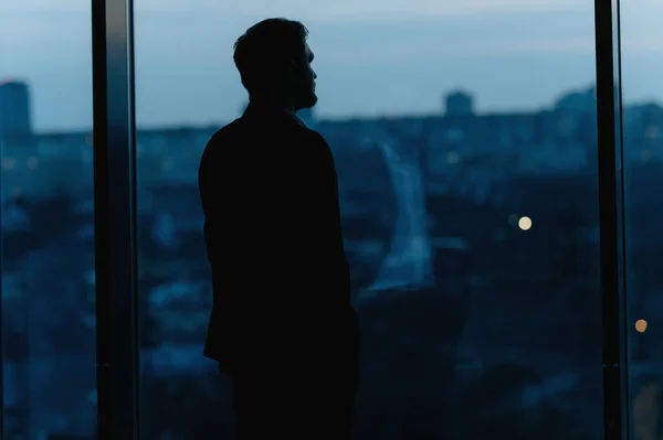 Gece vakti yüksek katlı ofis penceresinden dışarı bakan bir iş adamının silüeti.